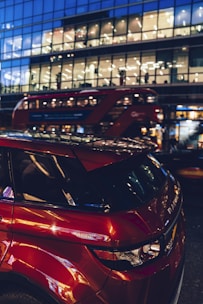 A modern building with large glass windows is illuminated against the evening sky, reflecting city lights and activity inside. A red double-decker bus moves past, adding a sense of urban life and motion. A sleek red Range Rover is parked in the foreground, highlighted by the ambient light.