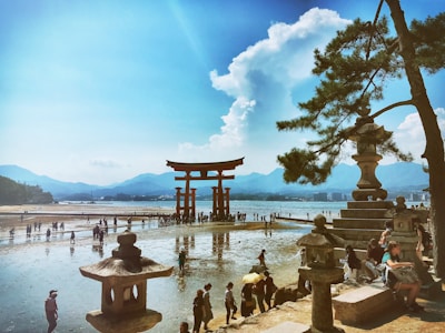 A scenic view of a large torii gate standing in the water during low tide, surrounded by many tourists walking on the wet sand. In the foreground, there are traditional stone lanterns and pine trees, with people relaxing and taking in the view. The backdrop features a range of blue mountains under a bright sky with fluffy clouds.
