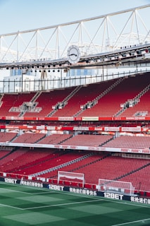 An empty football stadium with red seats, featuring advertisements along the perimeter of the field. The stadium has a large clock mounted on the upper structure, which is made of metal supports. The field is neatly mowed with clear white lines marking the boundaries.