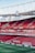 An empty football stadium with red seats, featuring advertisements along the perimeter of the field. The stadium has a large clock mounted on the upper structure, which is made of metal supports. The field is neatly mowed with clear white lines marking the boundaries.