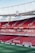 An empty football stadium with red seats, featuring advertisements along the perimeter of the field. The stadium has a large clock mounted on the upper structure, which is made of metal supports. The field is neatly mowed with clear white lines marking the boundaries.
