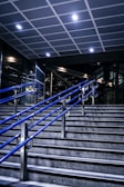 A staircase featuring blue metal handrails stretches upward in a modern indoor setting. Overhead, a tiled ceiling with recessed lighting illuminates the area. The surrounding walls and structures showcase a sleek, industrial design with glass and metal elements.