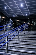 A staircase featuring blue metal handrails stretches upward in a modern indoor setting. Overhead, a tiled ceiling with recessed lighting illuminates the area. The surrounding walls and structures showcase a sleek, industrial design with glass and metal elements.