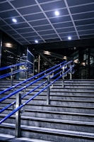 A staircase featuring blue metal handrails stretches upward in a modern indoor setting. Overhead, a tiled ceiling with recessed lighting illuminates the area. The surrounding walls and structures showcase a sleek, industrial design with glass and metal elements.