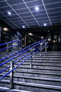 A staircase featuring blue metal handrails stretches upward in a modern indoor setting. Overhead, a tiled ceiling with recessed lighting illuminates the area. The surrounding walls and structures showcase a sleek, industrial design with glass and metal elements.