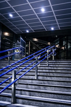 A staircase featuring blue metal handrails stretches upward in a modern indoor setting. Overhead, a tiled ceiling with recessed lighting illuminates the area. The surrounding walls and structures showcase a sleek, industrial design with glass and metal elements.