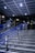 A staircase featuring blue metal handrails stretches upward in a modern indoor setting. Overhead, a tiled ceiling with recessed lighting illuminates the area. The surrounding walls and structures showcase a sleek, industrial design with glass and metal elements.