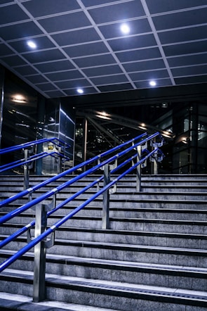 A staircase featuring blue metal handrails stretches upward in a modern indoor setting. Overhead, a tiled ceiling with recessed lighting illuminates the area. The surrounding walls and structures showcase a sleek, industrial design with glass and metal elements.