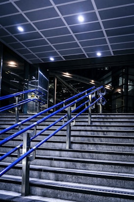 A staircase featuring blue metal handrails stretches upward in a modern indoor setting. Overhead, a tiled ceiling with recessed lighting illuminates the area. The surrounding walls and structures showcase a sleek, industrial design with glass and metal elements.