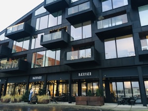 A modern building with large glass windows and balconies decorated with string lights. The structure features clean lines and a contemporary design, with a restaurant or cafe at the ground level named 'N&AElig;RV&AElig;R'. Some greenery in pots can be seen near the entrance, and tables with chairs are arranged outside. A person is visible near the entrance.