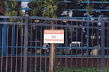 A metal fence with vertical bars surrounds a property. Attached to the fence is a sign with red and white lettering that reads 'Private Property No Trespassing.' Behind the fence, a tree and some greenery are visible, along with the reflection of a building's windows.