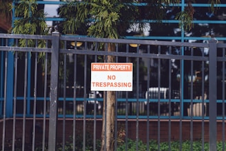A metal fence with vertical bars surrounds a property. Attached to the fence is a sign with red and white lettering that reads 'Private Property No Trespassing.' Behind the fence, a tree and some greenery are visible, along with the reflection of a building's windows.