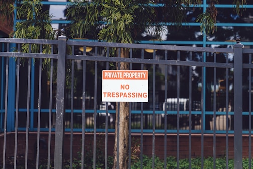 A metal fence with vertical bars surrounds a property. Attached to the fence is a sign with red and white lettering that reads 'Private Property No Trespassing.' Behind the fence, a tree and some greenery are visible, along with the reflection of a building's windows.