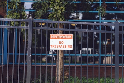 A metal fence with vertical bars surrounds a property. Attached to the fence is a sign with red and white lettering that reads 'Private Property No Trespassing.' Behind the fence, a tree and some greenery are visible, along with the reflection of a building's windows.
