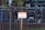 A metal fence with vertical bars surrounds a property. Attached to the fence is a sign with red and white lettering that reads 'Private Property No Trespassing.' Behind the fence, a tree and some greenery are visible, along with the reflection of a building's windows.