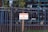 A metal fence with vertical bars surrounds a property. Attached to the fence is a sign with red and white lettering that reads 'Private Property No Trespassing.' Behind the fence, a tree and some greenery are visible, along with the reflection of a building's windows.
