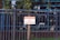 A metal fence with vertical bars surrounds a property. Attached to the fence is a sign with red and white lettering that reads 'Private Property No Trespassing.' Behind the fence, a tree and some greenery are visible, along with the reflection of a building's windows.