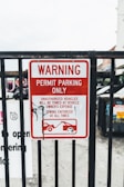 A metal fence with a red and white warning sign stating 'Permit Parking Only.' It indicates unauthorized vehicles will be towed at the owner's expense, with towing enforced at all times. There is a graphic of a tow truck and a car beneath the text. Some graffiti is visible on the sign.