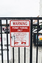 A metal fence with a red and white warning sign stating 'Permit Parking Only.' It indicates unauthorized vehicles will be towed at the owner's expense, with towing enforced at all times. There is a graphic of a tow truck and a car beneath the text. Some graffiti is visible on the sign.