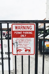 A metal fence with a red and white warning sign stating 'Permit Parking Only.' It indicates unauthorized vehicles will be towed at the owner's expense, with towing enforced at all times. There is a graphic of a tow truck and a car beneath the text. Some graffiti is visible on the sign.