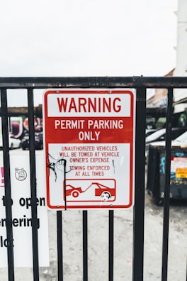 A metal fence with a red and white warning sign stating 'Permit Parking Only.' It indicates unauthorized vehicles will be towed at the owner's expense, with towing enforced at all times. There is a graphic of a tow truck and a car beneath the text. Some graffiti is visible on the sign.