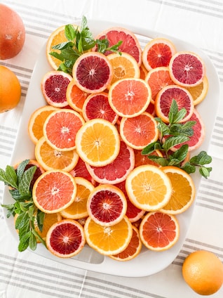 A white platter is filled with a variety of sliced citrus fruits, including oranges, grapefruits, and blood oranges. Fresh mint leaves are arranged on top for garnish. The colors of the fruit range from bright orange to deep red, creating a vibrant and refreshing appearance. The platter is placed on a white tablecloth with a subtle striped pattern. Whole oranges are visible in the background.