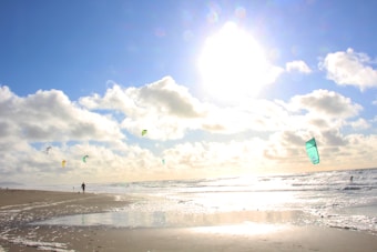 A sunny beach scene with a clear blue sky dotted with fluffy white clouds. Several kites are flying, with people engaging in kite surfing on the ocean. The sun casts a bright light over the water, creating a reflective shimmer on the wet sand.