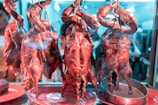 Several roasted ducks are hanging by hooks in a glass display, showcasing their shiny and crispy skin. The setting appears to be a food market or a restaurant with reflections visible on the glass.