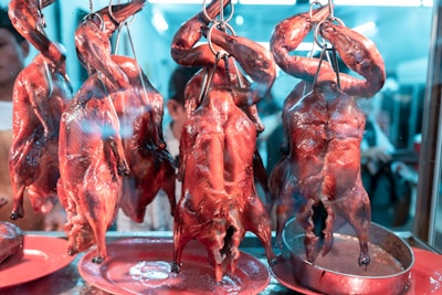 Several roasted ducks are hanging by hooks in a glass display, showcasing their shiny and crispy skin. The setting appears to be a food market or a restaurant with reflections visible on the glass.