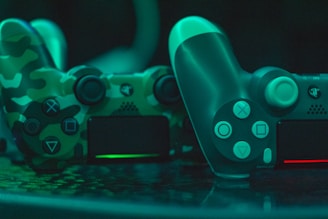 Two video game controllers are positioned closely together. One has a camouflage pattern with shades of green and black, while the other is solid in design with a teal tint. The controllers are illuminated with a green ambient light, creating a reflective effect on the surface they are placed on.