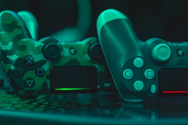 Two video game controllers are positioned closely together. One has a camouflage pattern with shades of green and black, while the other is solid in design with a teal tint. The controllers are illuminated with a green ambient light, creating a reflective effect on the surface they are placed on.