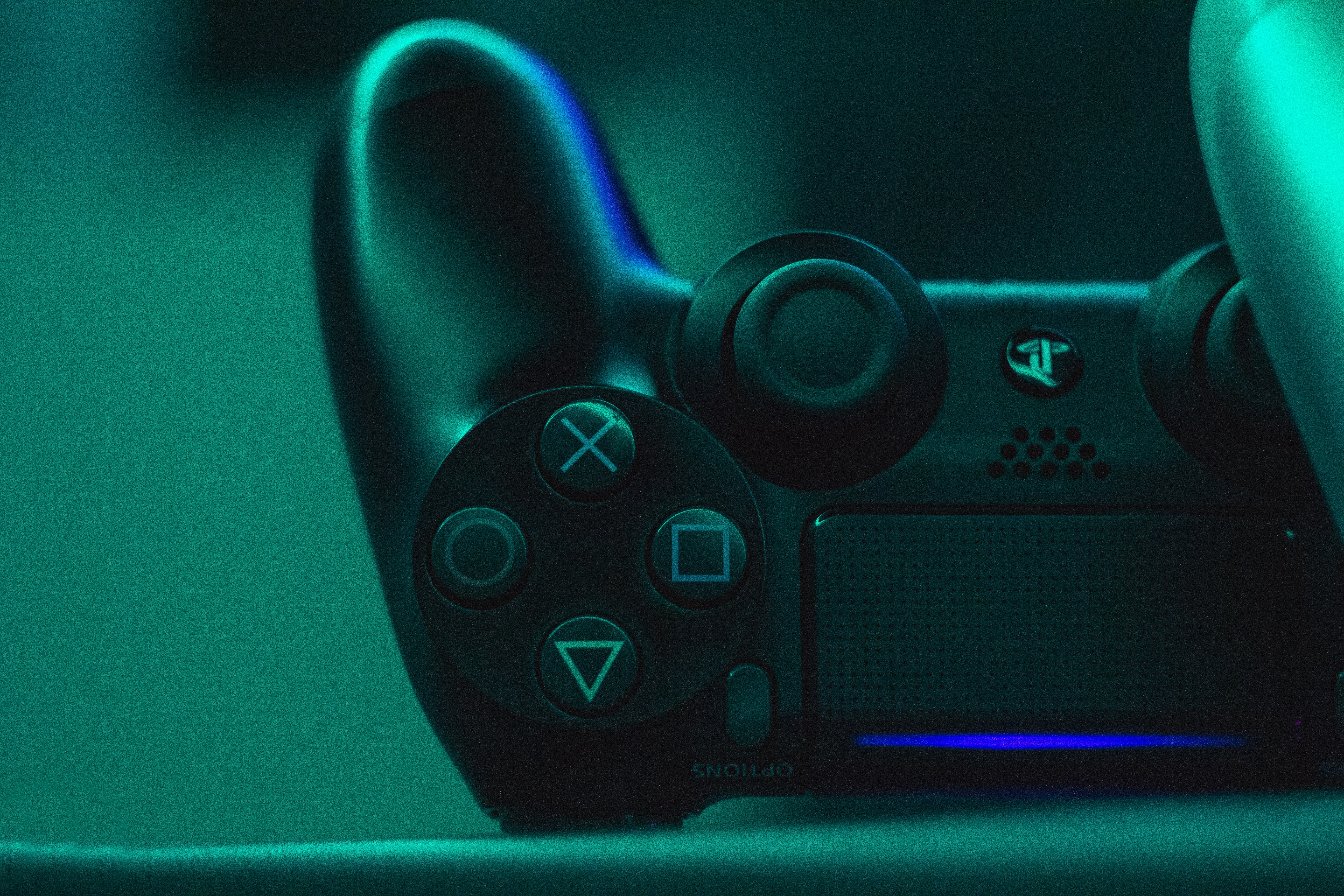 A close-up view of a black video game controller illuminated by a teal light, showcasing its buttons and analog sticks.