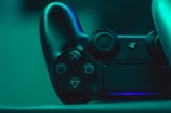 A close-up view of a black video game controller illuminated by a teal light, showcasing its buttons and analog sticks.