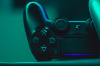 A close-up view of a black video game controller illuminated by a teal light, showcasing its buttons and analog sticks.