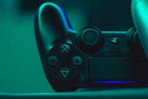 A close-up view of a black video game controller illuminated by a teal light, showcasing its buttons and analog sticks.