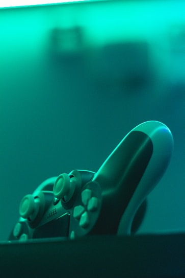 A close-up shot of a video game controller with focus on the joysticks and buttons. The lighting creates a teal ambiance, giving the scene a futuristic atmosphere.