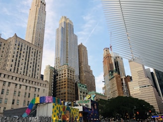 A bustling urban scene featuring a skyline filled with tall skyscrapers of varying architectural styles. These towering buildings are a mix of both modern glass structures and older, ornate constructions. In the foreground, there is a colorful, artistic installation with vibrant patterns and murals. A large white, rib-like structure is partially visible on the right. The scene conveys a sense of a lively, dynamic city environment.