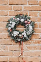 A festive holiday wreath decorated with silver, red, and white ornaments along with evergreen branches. It is hanging on a brick wall, with some artificial snow on the ornaments and greenery. A ribbon in red and white hangs from the bottom of the wreath.