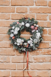 A festive holiday wreath decorated with silver, red, and white ornaments along with evergreen branches. It is hanging on a brick wall, with some artificial snow on the ornaments and greenery. A ribbon in red and white hangs from the bottom of the wreath.
