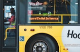 A yellow city tour bus has a digital sign indicating it is not in service. A person in a reflective vest can be seen inside the bus, near the door. Various signs about accessibility and passenger instructions are visible on the bus.
