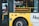 A yellow city tour bus has a digital sign indicating it is not in service. A person in a reflective vest can be seen inside the bus, near the door. Various signs about accessibility and passenger instructions are visible on the bus.