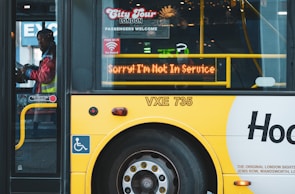 A yellow city tour bus has a digital sign indicating it is not in service. A person in a reflective vest can be seen inside the bus, near the door. Various signs about accessibility and passenger instructions are visible on the bus.