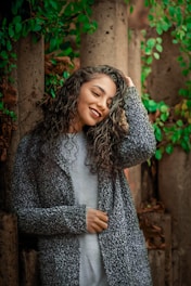 A person with long, curly hair and a light gray top stands in front of a backdrop with vertical stone columns and lush green foliage. The individual is smiling warmly and holding a hand to their head, exuding a sense of relaxed confidence.