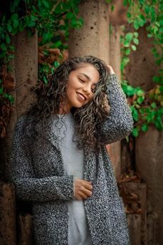 A person with long, curly hair and a light gray top stands in front of a backdrop with vertical stone columns and lush green foliage. The individual is smiling warmly and holding a hand to their head, exuding a sense of relaxed confidence.