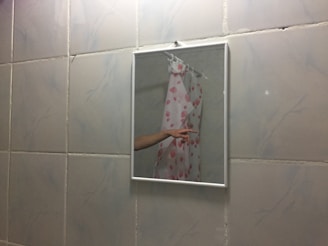 A bathroom mirror reflects part of a shower curtain with a pink seashell pattern and an extended arm pointing. The wall is tiled with white tiles featuring light grey marbling.