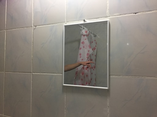 A bathroom mirror reflects part of a shower curtain with a pink seashell pattern and an extended arm pointing. The wall is tiled with white tiles featuring light grey marbling.