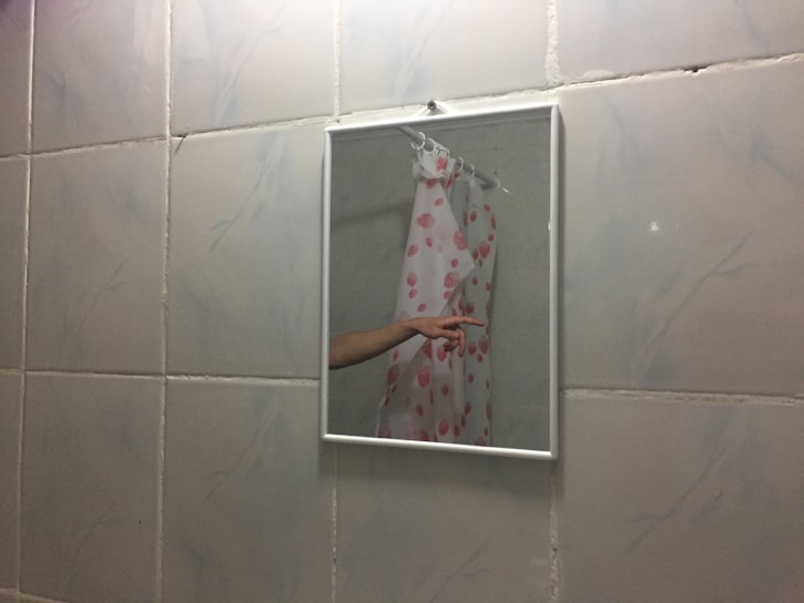 A bathroom mirror reflects part of a shower curtain with a pink seashell pattern and an extended arm pointing. The wall is tiled with white tiles featuring light grey marbling.