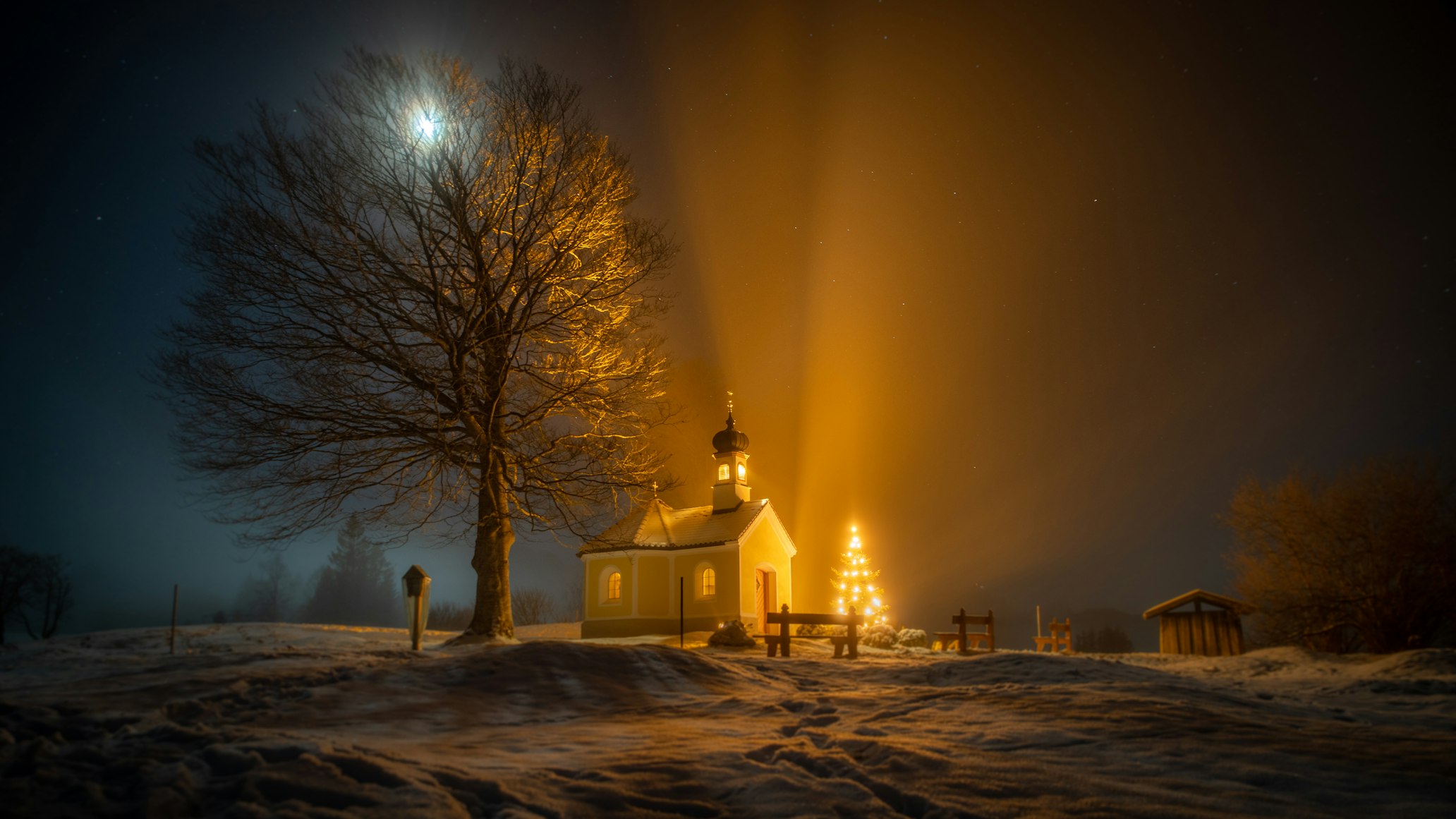 Nighttime photo of a small church in snowy landscape with glowing christmas tree in yard.