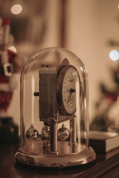 A vintage-style clock is encased in a glass dome with ornamental metal balls at its base. The design features a classic clock face with Roman numerals. The background is softly blurred, suggesting a warm, ambient setting.