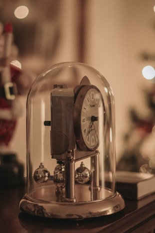 A vintage-style clock is encased in a glass dome with ornamental metal balls at its base. The design features a classic clock face with Roman numerals. The background is softly blurred, suggesting a warm, ambient setting.